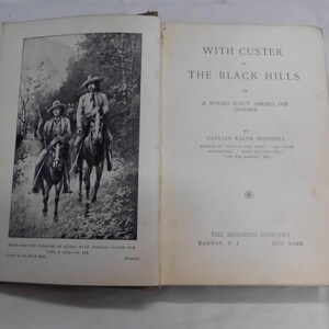 May include: Open book with the title "With Custer in the Black Hills" by Captain Ralph Bonehill. The left page features a black and white illustration of two men on horseback. The book is from The Mershon Company.