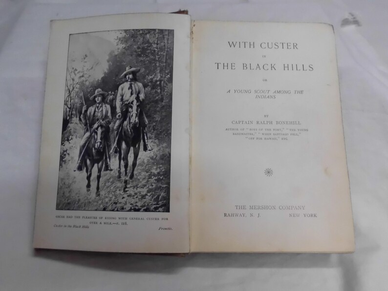 May include: Open book with the title "With Custer in the Black Hills" by Captain Ralph Bonehill. The left page features a black and white illustration of two men on horseback. The book is from The Mershon Company.