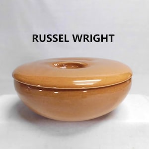 May include: A round, tan-colored ceramic bowl with a matching lid featuring a central circular indentation. The text RUSSEL WRIGHT is printed in black above the bowl. The piece has a smooth, glossy finish and a simple, curved silhouette.