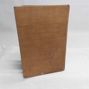 May include: A rectangular, brown, vintage book with a textured cover. The book is standing upright, with the spine facing the viewer. The cover has a slightly worn appearance, suggesting age and use. The book is set against a white background.