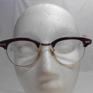 May include: Vintage eyeglasses with a dark burgundy upper frame and gold-colored lower frame. The glasses have a classic browline design and clear lenses. The frame features small gold accents on the temples.