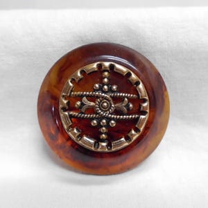 May include: A round, brown button with a decorative brass-colored center. The button has a marbled appearance with shades of amber and brown. The center features an ornate design with a symmetrical pattern.