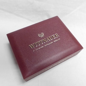 May include: A burgundy-colored Wittnauer watch box. The box has gold lettering that reads "WITTNAUER" and "A LONGINES - WITTNAUER PRODUCT". The box is square and has a gold trim.