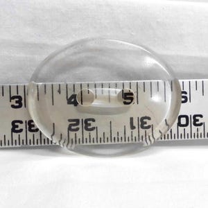 May include: A clear, oval-shaped button with two holes, approximately 1.5 inches in diameter. The button is placed on a white surface next to a ruler for size comparison. The button is made of a transparent material.