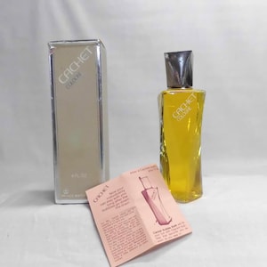 May include: A vintage bottle of Cachet Cologne with a silver-toned box and a small pamphlet. The cologne bottle is a unique shape with a silver cap and contains a yellow liquid. The box and bottle both display the Cachet logo.