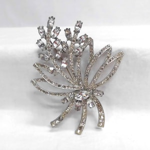 Op de afbeelding: Een zilverkleurige metalen broche in de vorm van een bloemtak met lintachtige lussen. Het sieraad is bezet met talloze heldere, gefacetteerde strass-steentjes in diverse formaten, waaronder markies- en ronde slijpvormen.