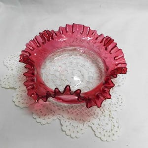 May include: A cranberry-colored glass bowl with a ruffled rim. The bowl sits on a white, lacy doily. The glass is transparent, and the ruffled edge adds a decorative touch to the piece. The bowl is likely vintage.