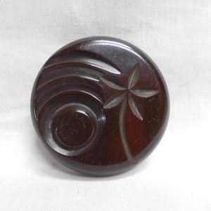 May include: A dark brown, circular button with carved designs. The button features a stylized palm tree and curved lines, possibly made of Bakelite or a similar material. The button is set against a white background.
