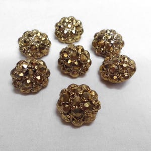 May include: Seven gold-colored, clustered buttons with a faceted, textured surface. The buttons are arranged on a white background. The buttons have a metallic sheen and a rounded shape, suitable for sewing or craft projects.