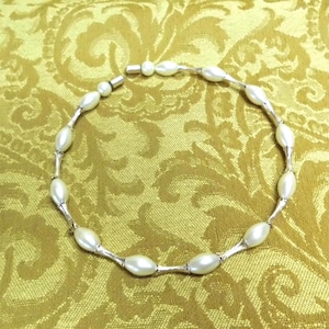 Puede incluir: Un collar con perlas blancas de forma ovalada unidas por barras de metal plateado. El collar se muestra sobre una tela dorada estampada.