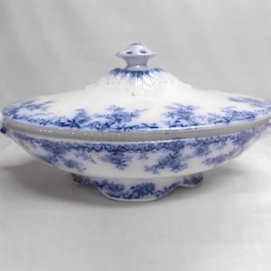 May include: An oval ceramic tureen with a lid, featuring a white base decorated with intricate blue floral patterns. The piece includes two side handles, a scalloped base, and a central knob on the lid, all adorned with matching blue designs.