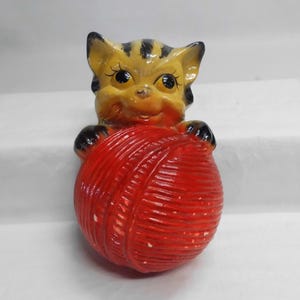 May include: A ceramic figurine of a cat holding a red ball of yarn. The cat has yellow fur with black stripes and black paws. The yarn ball is a vibrant red color.