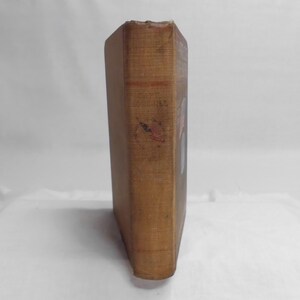 May include: A vintage book with a worn, tan cover. The spine shows signs of age and wear, with faded text and a small, red and black mark. The book's pages are not visible, suggesting a focus on the cover's aesthetic.