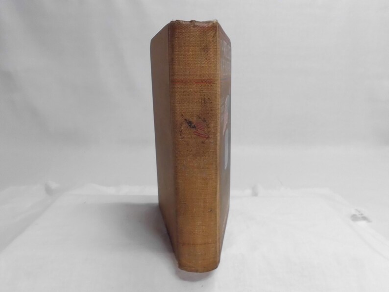 May include: A vintage book with a worn, tan cover. The spine shows signs of age and wear, with faded text and a small, red and black mark. The book's pages are not visible, suggesting a focus on the cover's aesthetic.