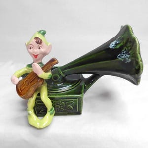 May include: A ceramic figurine of a green-clad elf playing a brown guitar, seated next to a dark green vintage-style phonograph with a large horn. The base of the phonograph features a molded floral relief pattern in a glossy glazed finish.