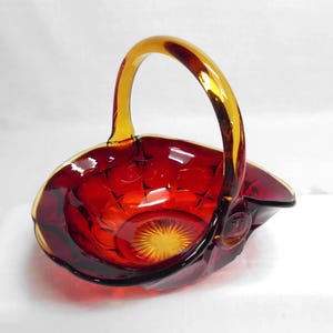 May include: A ruby red glass basket with a golden handle. The basket has a diamond pattern on the sides and a starburst design on the base. The handle is arched and attached to the rim of the basket.