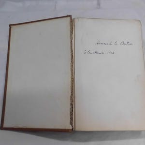 May include: An open book with aged, off-white pages. The left page is blank, while the right page features handwritten text: "Howard G. Bates. Christmas. 1903." The book's cover is a dark brown color.