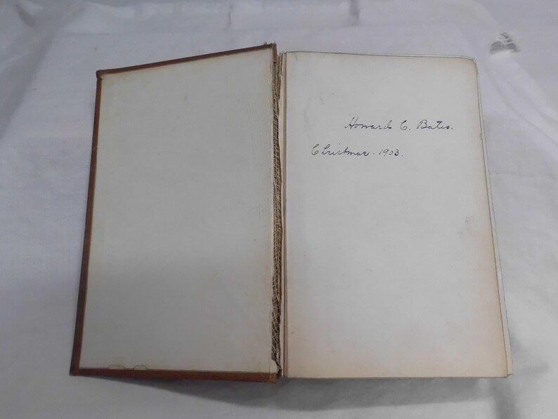 May include: An open book with aged, off-white pages. The left page is blank, while the right page features handwritten text: "Howard G. Bates. Christmas. 1903." The book's cover is a dark brown color.