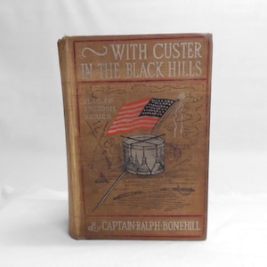 May include: Antique book titled "With Custer in the Black Hills" by Captain Ralph Bonehill. The cover features an illustration of the American flag, a drum, and the text "Flag of Freedom Series."