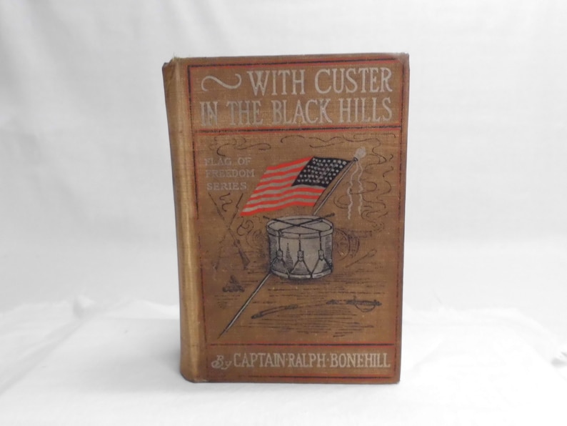 May include: Antique book titled "With Custer in the Black Hills" by Captain Ralph Bonehill. The cover features an illustration of the American flag, a drum, and the text "Flag of Freedom Series."