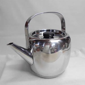May include: A polished stainless steel teapot with a rounded body, a curved spout, and a high arched handle. The lid features a small, circular black knob centered on top. The reflective surface of the metal shows a distorted view of the surroundings.