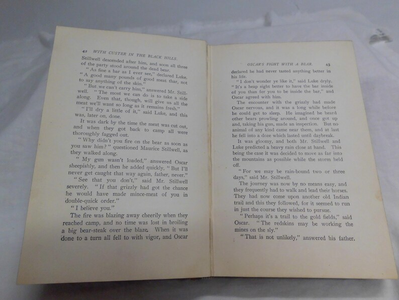 May include: Open book with text on aged paper. The left page has the title "WITH CESTER IN THE BLACK HILLS". The right page has the title "DICAR'S FIGHT WITH A BEAR".