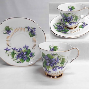 May include: A white teacup and saucer set with a floral violet design. The teacup has a gold base and handle, and the saucer has a gold rim. The violet flowers are purple and green.