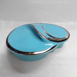 May include: A bright turquoise oval-shaped container with a smooth, glossy finish and a metallic silver trim along the edges. The lid features a curved, recessed design that creates a distinct wave-like pattern across the top surface of the object.