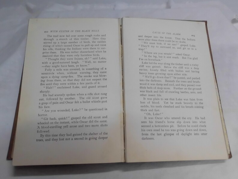 May include: Open book with text on both pages. The pages are yellowed with age, and the text is in black. The book is open to a chapter titled "With Custer in the Black Hills" and "Cause of the Alarm."