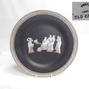 May include: A black ceramic plate with a gold rim and a white and blue patterned border. The plate features a relief of classical figures in white and pink. The text "PRATT FENTON OLD GREEK" is visible.