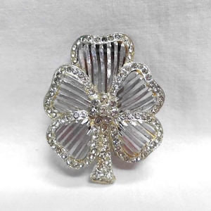 May include: A silver-toned clover brooch with clear crystal accents. The brooch features a three-leaf clover design with a central cluster of crystals and a border of smaller crystals. The leaves have a ribbed texture.