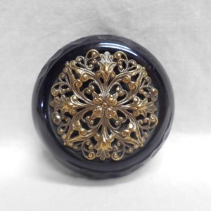 May include: A round, black button featuring a central, intricate, gold-toned metal filigree overlay. The black base has a smooth, glossy finish with subtle, scalloped edges around the perimeter, contrasting with the detailed, ornate metalwork.