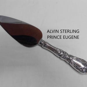 May include: A silver cake server with a polished, triangular blade and an ornate, embossed handle featuring intricate floral and scrollwork details. The text ALVIN STERLING PRINCE EUGENE is printed clearly next to the handle of the utensil.