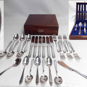 May include: A complete silver-toned flatware set, including spoons, forks, knives, and serving pieces. The set is arranged on a white surface, with a wooden storage box in the background. Some pieces are displayed inside a blue velvet-lined box.