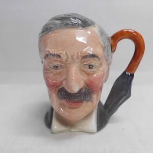 May include: A ceramic mug shaped like a man's head, featuring a gray-haired man with a mustache and a red-tinged face. The handle is a brown umbrella handle. The mug is a unique collectible.