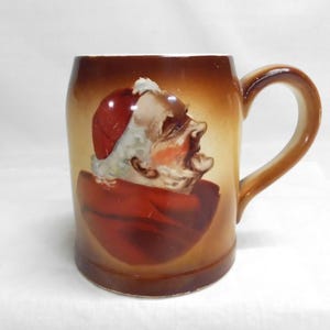 May include: A ceramic mug with a brown handle and a painted image of a man in a red hat and cloak. The mug has a brown and tan gradient background. The man's face is detailed with white hair and a red face.