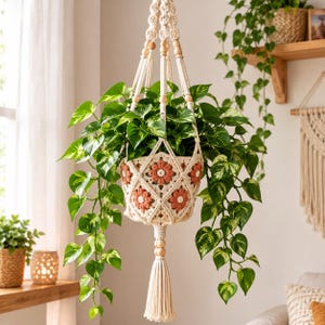 May include: A cream-colored macrame plant hanger featuring a crocheted basket with terracotta-colored flower motifs. The hanger includes natural wooden beads, a long bottom tassel, and holds a lush green pothos plant with variegated leaves.