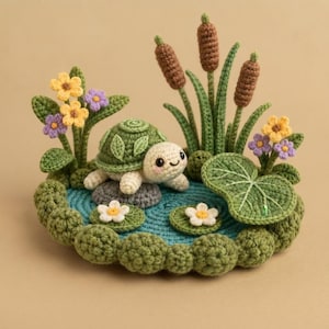 Puede incluir: Una figura de tortuga tejida a ganchillo sobre una piedra gris en un estanque circular azul. La escena incluye espadañas verdes, hojas de nenúfar y pequeñas flores amarillas y moradas. La tortuga tiene un caparazón verde y un cuerpo color crema.