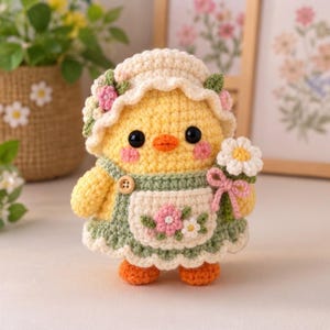 May include: A yellow crocheted chick doll wearing a sage green apron with a floral pattern, a matching bonnet adorned with small pink and green flowers, and holding a white daisy with a pink bow. The chick has black eyes and orange feet.