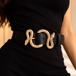May include: A black belt featuring a gold-toned metal buckle shaped like a coiled snake. The belt strap has a smooth, matte texture and is secured with a matching black leather-like loop. The snake buckle displays a detailed, textured scale pattern.