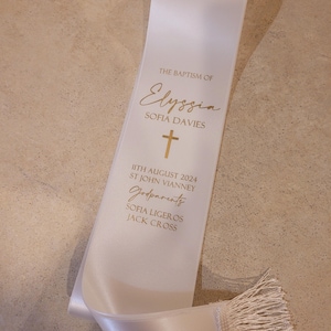 May include: A white satin sash featuring gold printed text that reads THE BAPTISM OF Elyssia SOFIA DAVIES, a gold cross icon, the date 11TH AUGUST 2024, ST JOHN VIANNEY, the word Godparents, and the names SOFIA LIGEROS and JACK CROSS, with fringe.