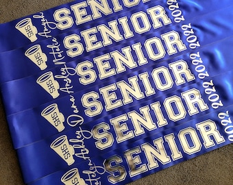 Può includere: Sei nastri di laurea blu e argentati con la parola "SENIOR" stampata in argento su ogni nastro. I nastri hanno anche l'anno "2022" stampato su di essi. Ogni nastro ha un piccolo megafono argentato con le lettere "SHS" stampate su di esso.