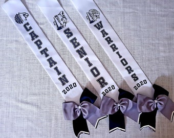May include: Four white sashes with black text and images. The sashes read "Captain 2020", "Senior 2020", "Warriors 2020", and an image of a Native American warrior. Each sash has a black and gray bow.