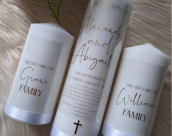May include: Three white pillar candles with gold lettering. The center candle reads "Alexande and Abigail" with wedding details. The other candles read "Two Become One" and a family name.