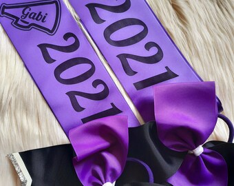 May include: Two purple sashes with black text and a megaphone graphic. The sashes say "Gabi 2021" and "Melody 2021". Two purple and black hair bows are also included.