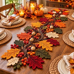 May include: A decorative autumn-themed table runner featuring crocheted oak leaves in shades of red, orange, yellow, green, and cream, accented with small crocheted berries and acorns, arranged in a winding vine pattern along the centre of a table.