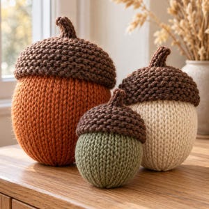 May include: Three handcrafted knitted acorn-shaped decorations in varying sizes. The largest is orange, the medium is cream, and the smallest is sage green. Each features a dark brown textured cap with a small stem on top.