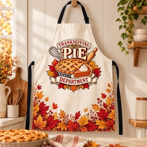 May include: A cream-colored apron featuring a central graphic of a lattice-top pie, a whisk, and a rolling pin with the text "THANKSGIVING PIE DEPARTMENT." The bottom border is decorated with a vibrant pattern of red, orange, and yellow autumn leaves.