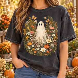 Pode incluir: Uma t-shirt de manga curta cinza-carvão com um gráfico central de um fantasma branco rodeado por uma coroa vibrante de flores de outono coloridas, folhagem verde e várias borboletas cor de laranja, impressa na parte da frente.