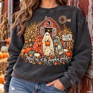 Pode incluir: Uma camisola de gola redonda cinzento-carvão com um gráfico de três fantasmas a segurar abóboras em frente a um celeiro vermelho. O design inclui pés de milho, um moinho de vento e o texto "Moonlit Pumpkin Farm" e "Meet me at the pumpkin patch."